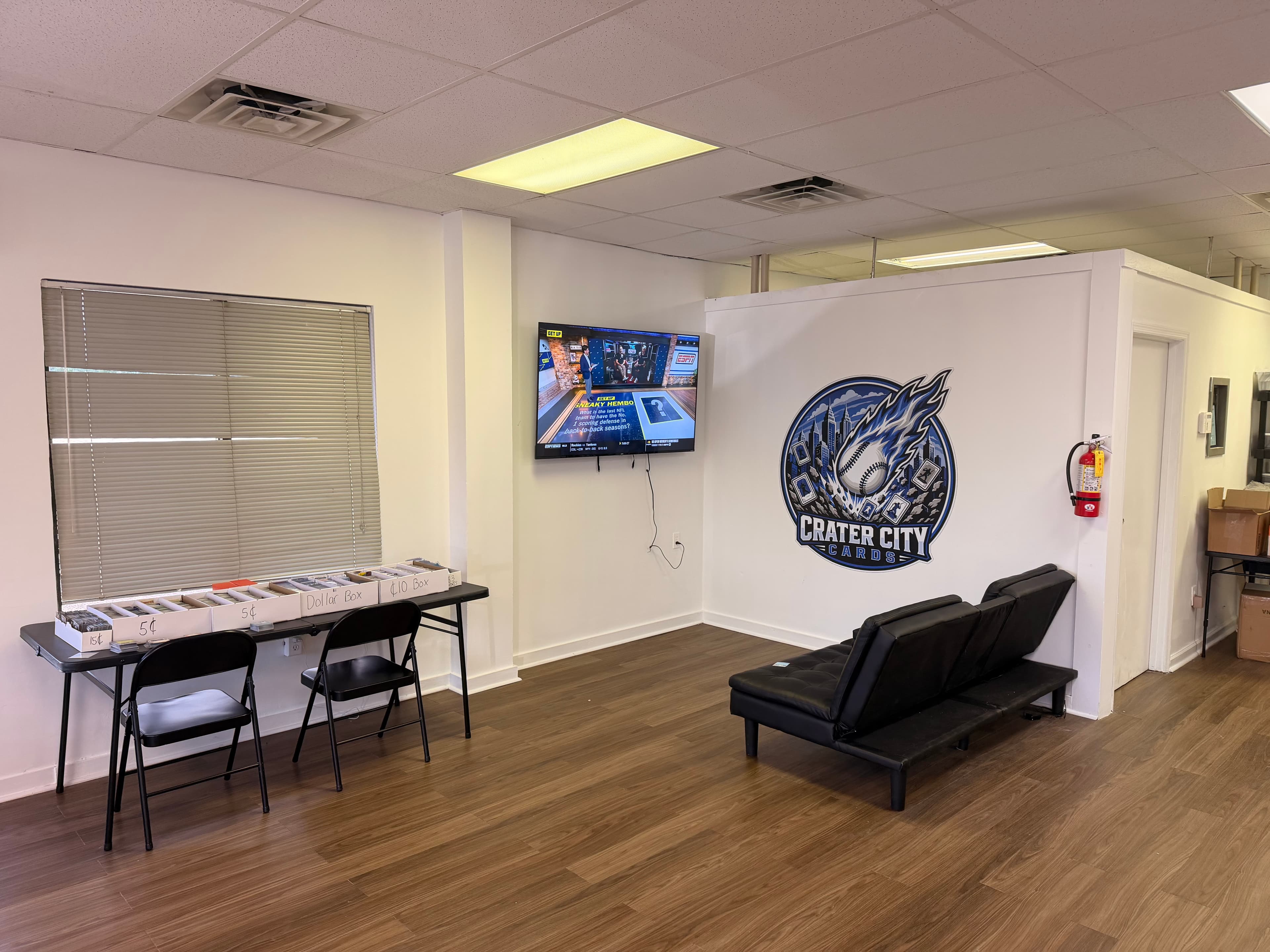 Crater City Cards shop interior with card boxes, a wall-mounted TV, and black sofa.